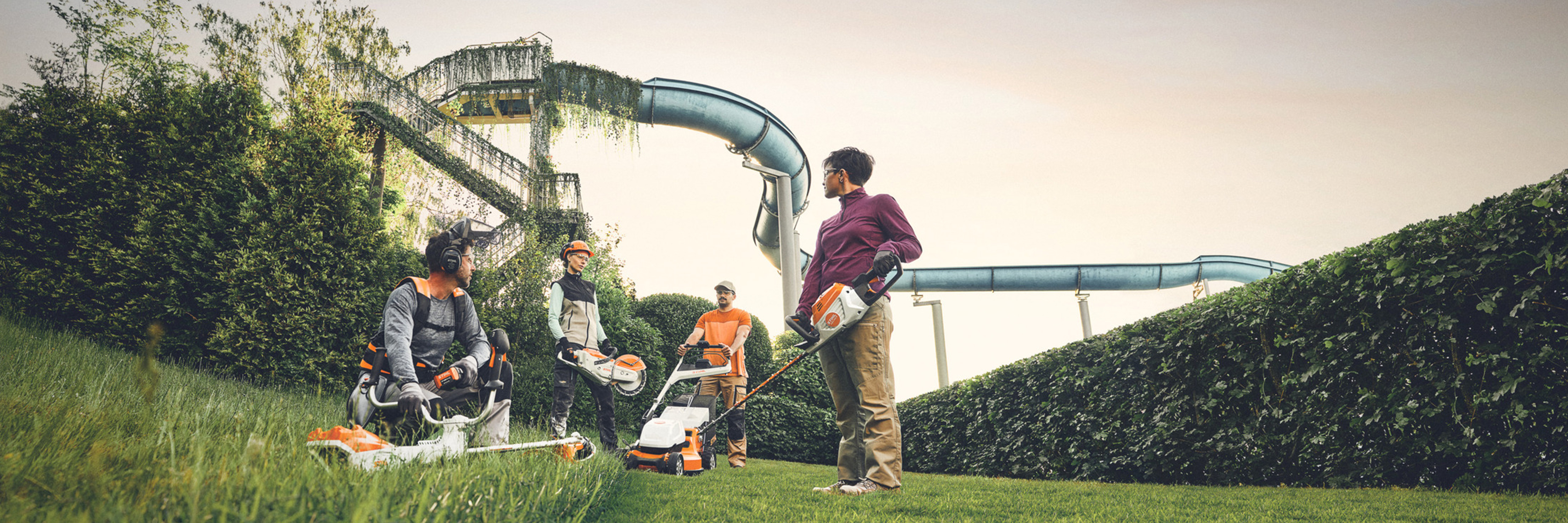 Dans un espace vert avec une grande haie et des toboggans aquatiques en arrière-plan, plusieurs personnes travaillent avec des outils de jardinage STIHL. Elles sont équipées de débroussailleuses, de tondeuses à gazon et d'équipements de protection.