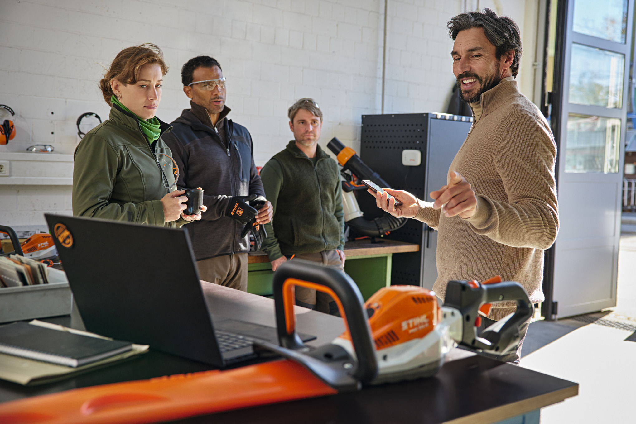 Vier personen staan in een werkplaats en een van hen legt de STIHL connected app op een smartphone uit