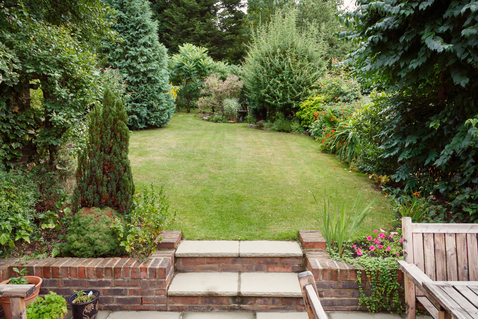 Petit jardin avec patio, mobilier de jardin et pots de plantes à l’avant-plan et pelouse avec arbustes à l'arrière.