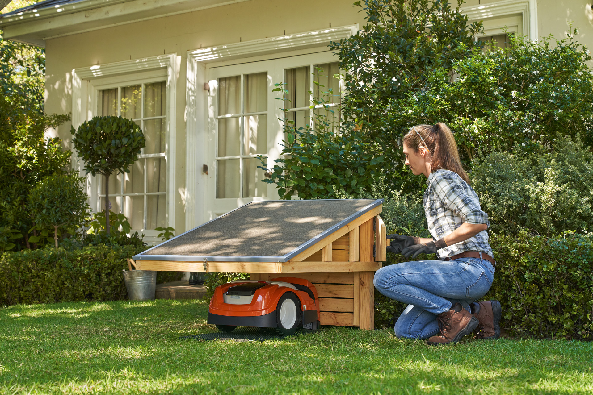 Une femme portant des lunettes de sécurité et des gants de travail s’agenouille sur la pelouse à côté d’un abri STIHL iMOW® où se trouve un robot de tonte STIHL iMOW® et ouvre le compartiment de rangement