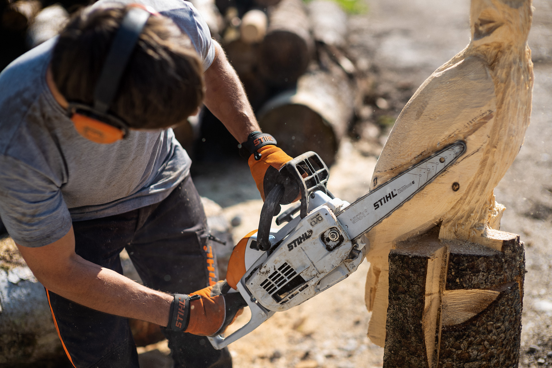 Un homme équipé de protections auditives et des gants utilisant une tronçonneuse STIHL MS 193 C-E pour travailler sur une sculpture en bois d’un oiseau partiellement terminée
