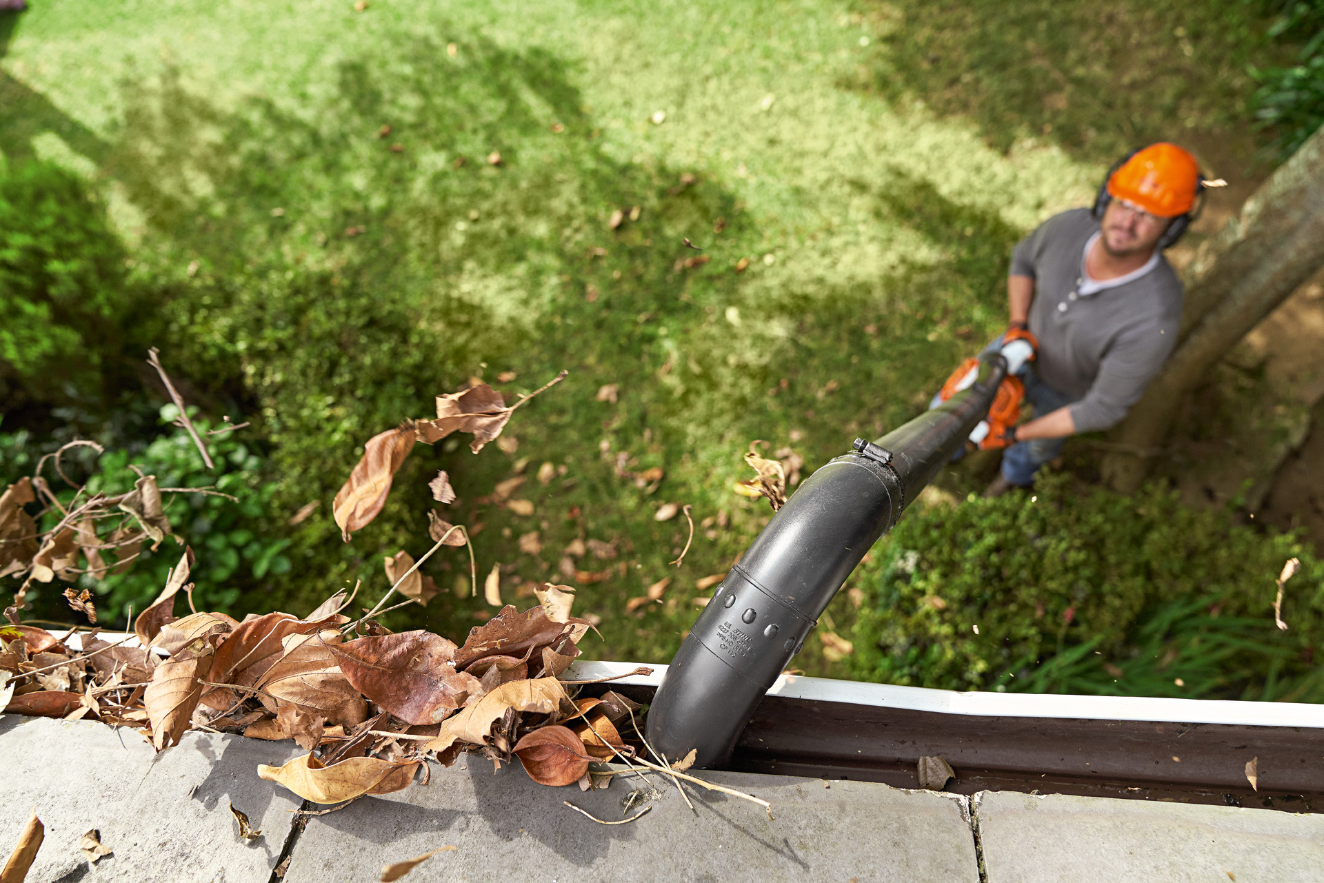 Een dakgoot vol bladeren wordt gereinigd door een man op de grond met een STIHL bladblazer en dakgootreinigingsset