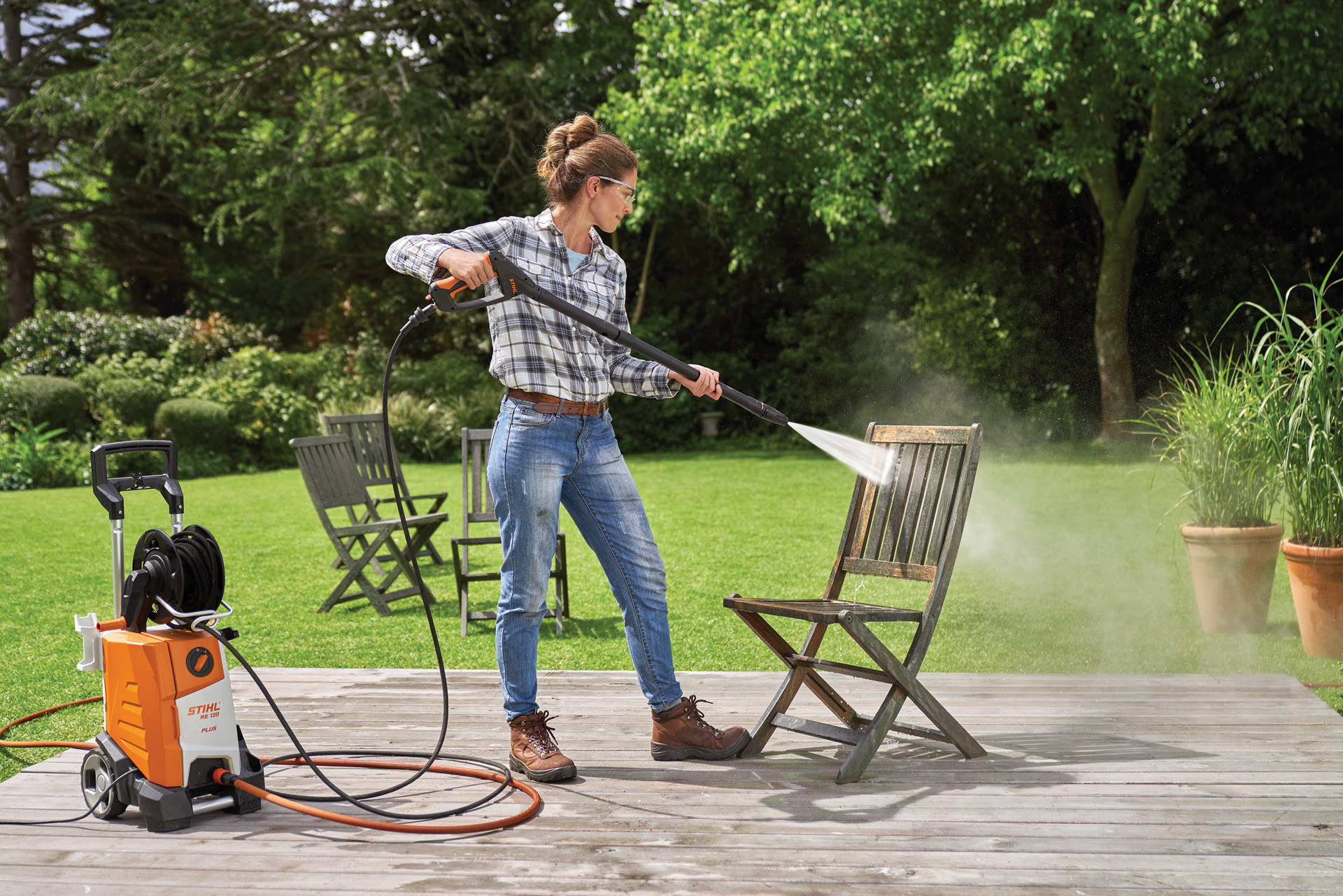 Een vrouw die een STIHL RE 130 Plus hogedrukreiniger gebruikt om een houten tuinstoel op een terras schoon te maken