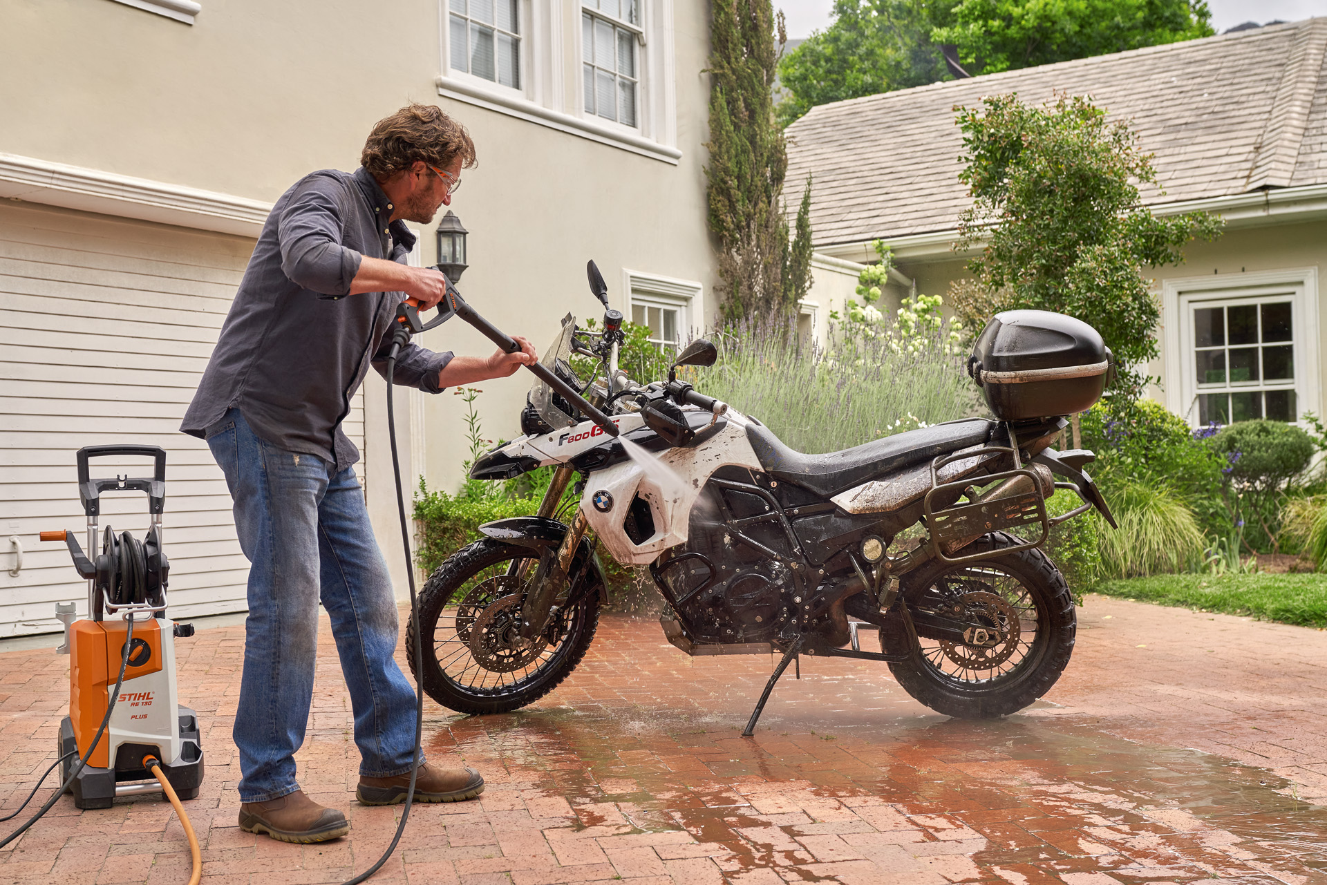 Een man gebruikt een STIHL RE 130 Plus hogedrukreiniger om thuis een witte motor op de oprit te wassen.