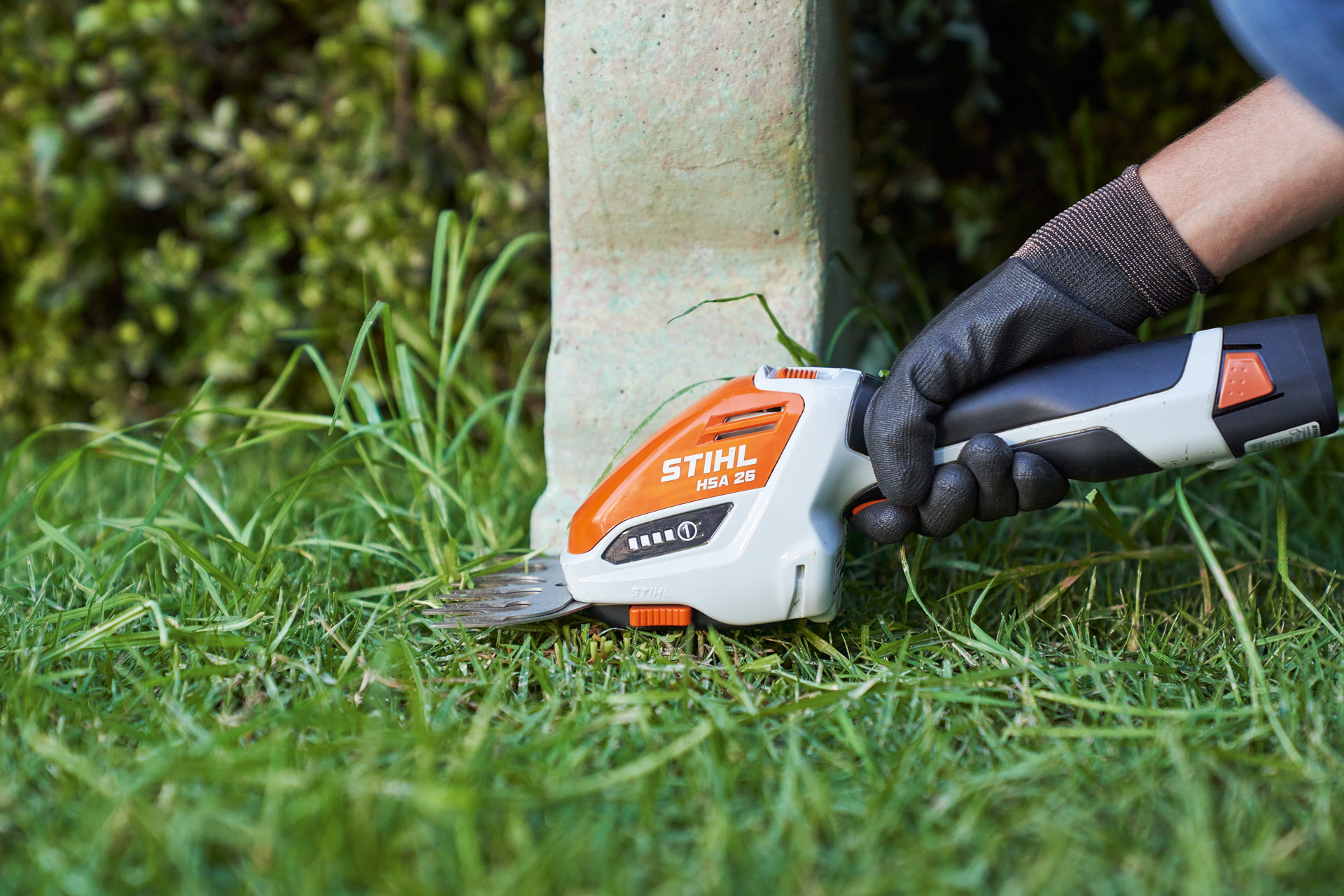 Close-up: persoon met veiligheidshandschoen trimt gazonrand met STIHL accugras- en buxusschaar HSA 26 rond een bank