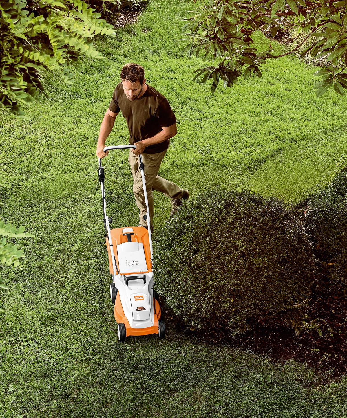 Vue plongeante : Homme tondant du gazon autour d'un petit arbre avec la tondeuse à batterie STIHL RMA 235.