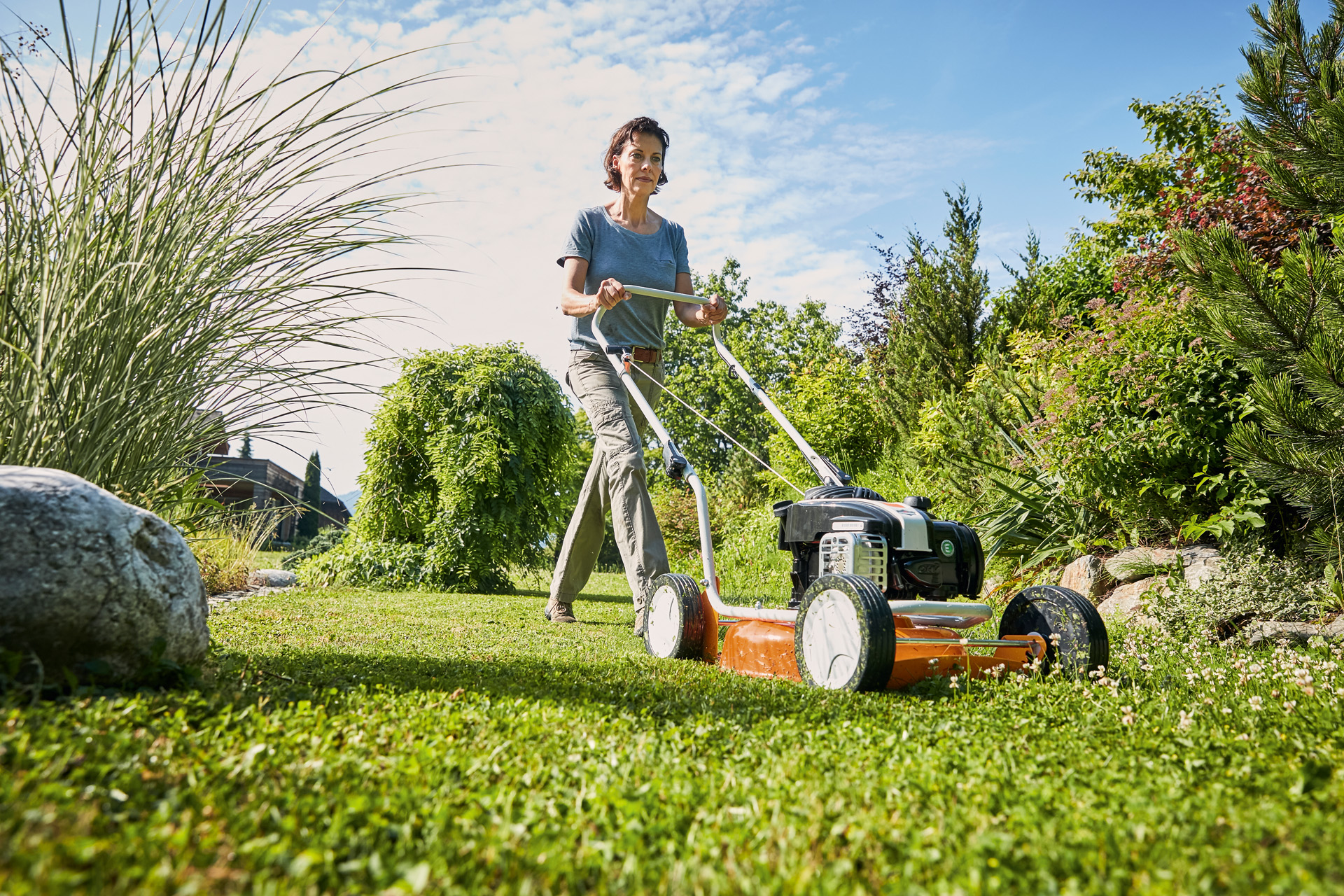 Une femme utilisant une tondeuse thermique STIHL RM 2 R sur une pelouse bordée de buissons et d’herbes