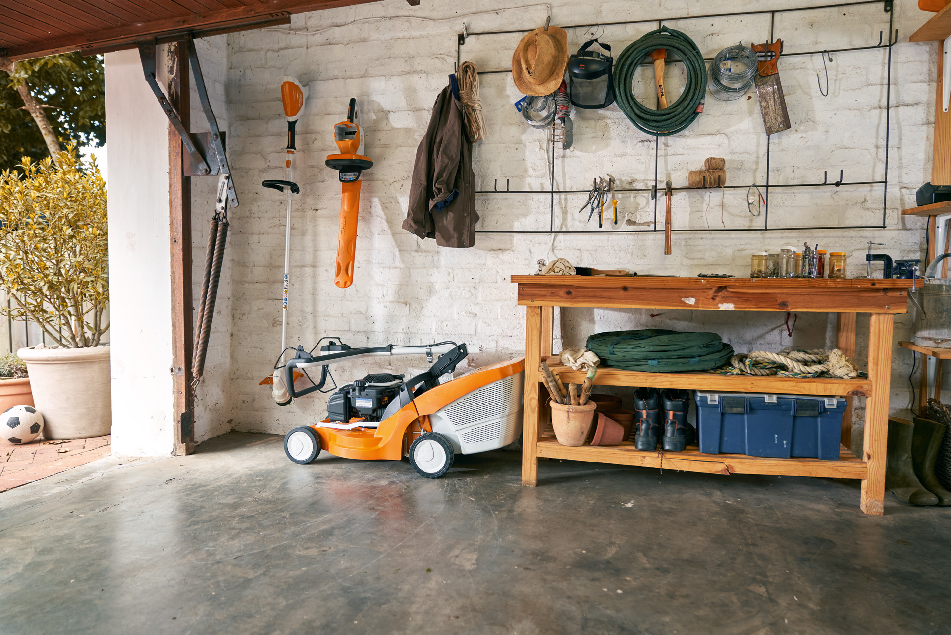 Een vrouw met handschoenen klapt de duwbeugel van een grasmaaier om in een garage met een werkbank op de achtergrond.