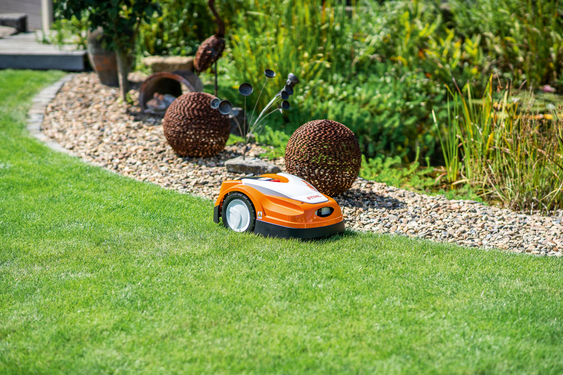 Une tondeuse robot STIHL iMOW® RMI 422 sur une pelouse à côté d’un lit de gravier et de sculptures de jardin