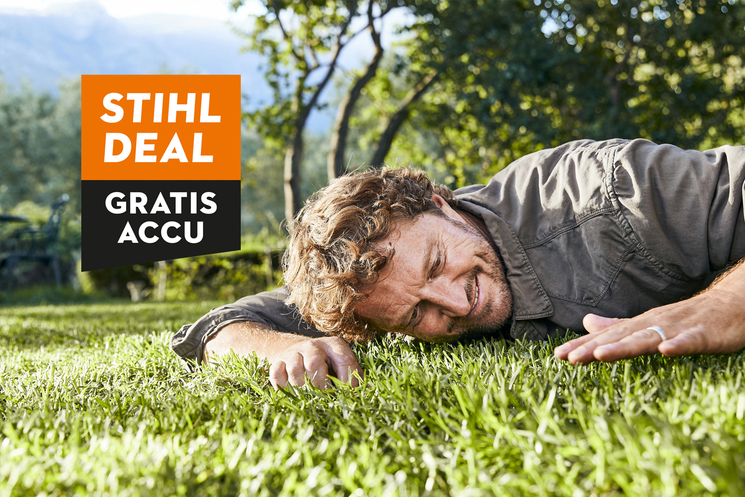 Een man ligt op de grond en aait blij het gemaaide gazon. Er is een oranje stoorelement over de actie met gratis AK 20 accu te zien. 
