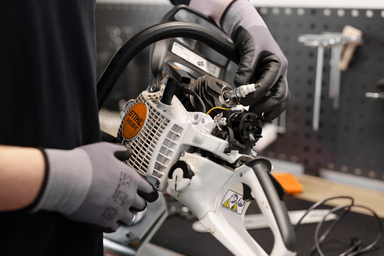 Dans un atelier, une personne portant des gants se tient au premier plan, tenant le moteur d'une tronçonneuse et changeant une bougie d'allumage.