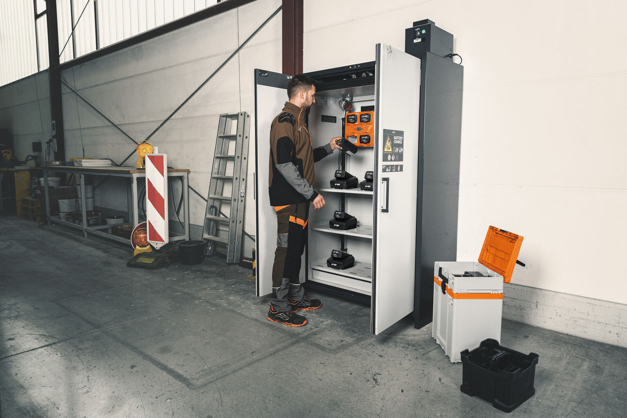Au premier plan, une femme blonde en tenue STIHL près de l’armoire de charge de batteries dans un atelier, à l’arrière-plan, un homme aux cheveux gris chargeant un véhicule.