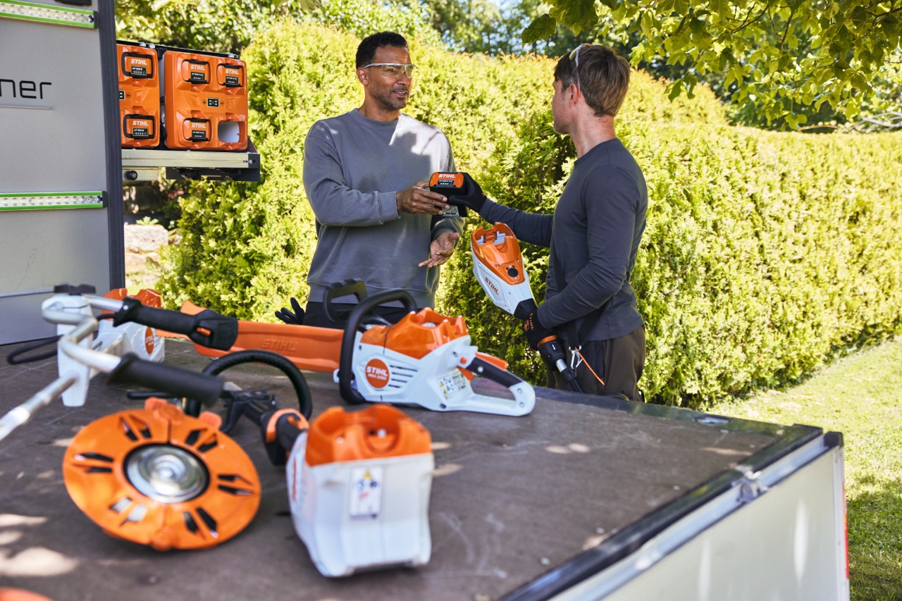 Een man geeft zijn collega een STIHL accu, ze bevinden zich allebei achter de laadvloer van een voertuig, waarop onder andere de bottTainer en een STIHL accukettingzaag en andere apparaten staan