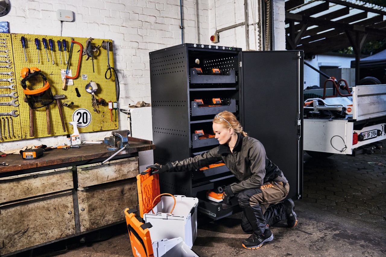 Vrouw in STIHL werkkleding voor een accu-oplaadkast en STIHL connected box, op de achtergrond een gereedschapswand 